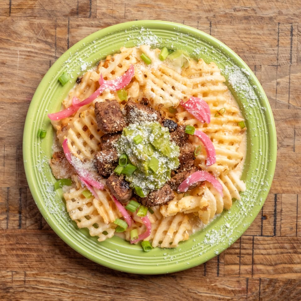 Waffle fries topped with steak, guacamole, pickled red onions, and green onions on a green plate.