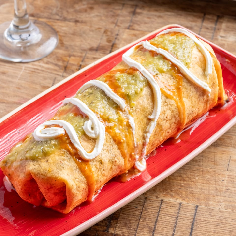 A large, golden-brown burrito topped with green salsa, red sauce, and white sour cream on a red plate.