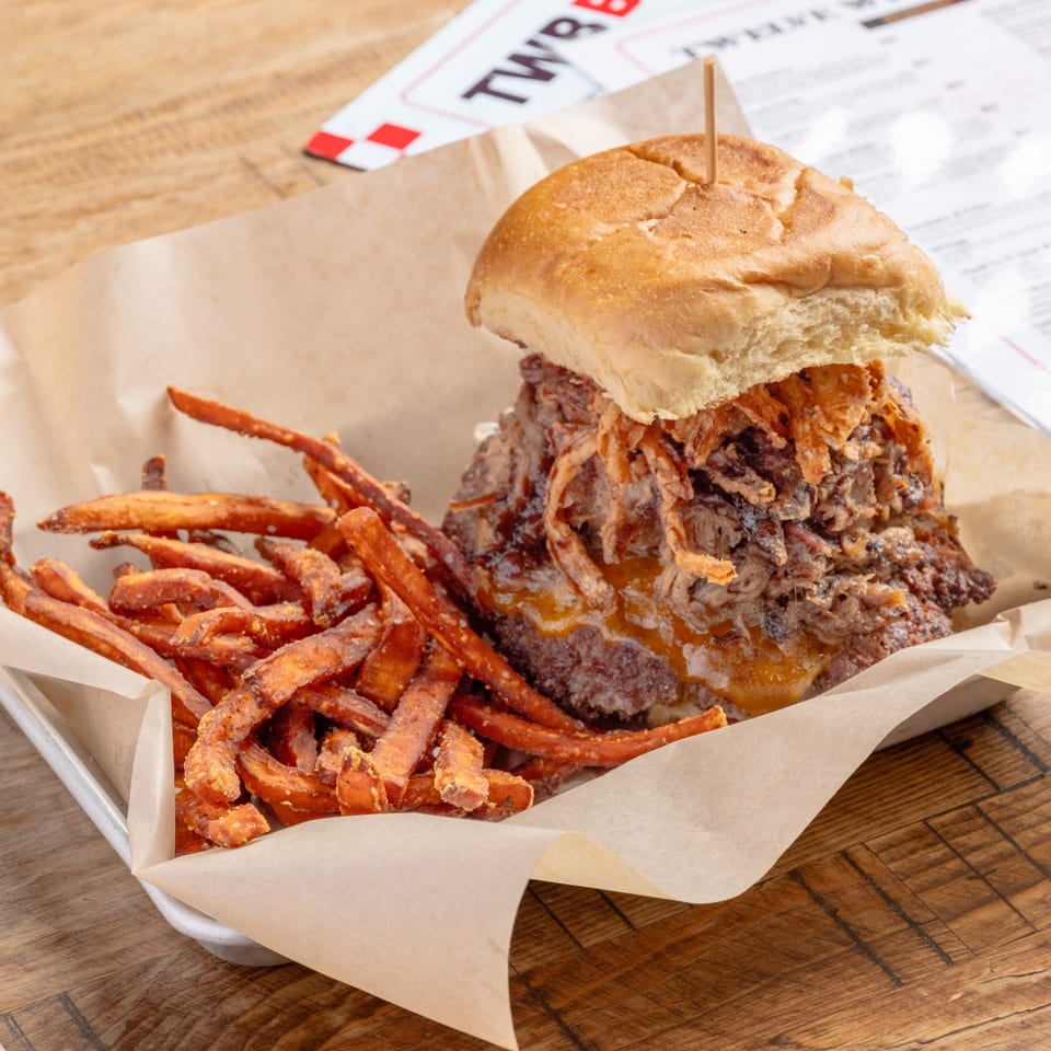 A large burger with pulled pork and crispy onions, served with sweet potato fries in a paper-lined tray.